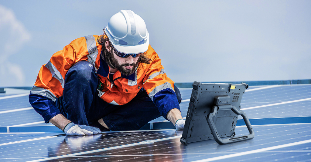 Utility field technician in high-visibility gear inspecting solar panels with a Getac G140 rugged tablet positioned nearby on the installation