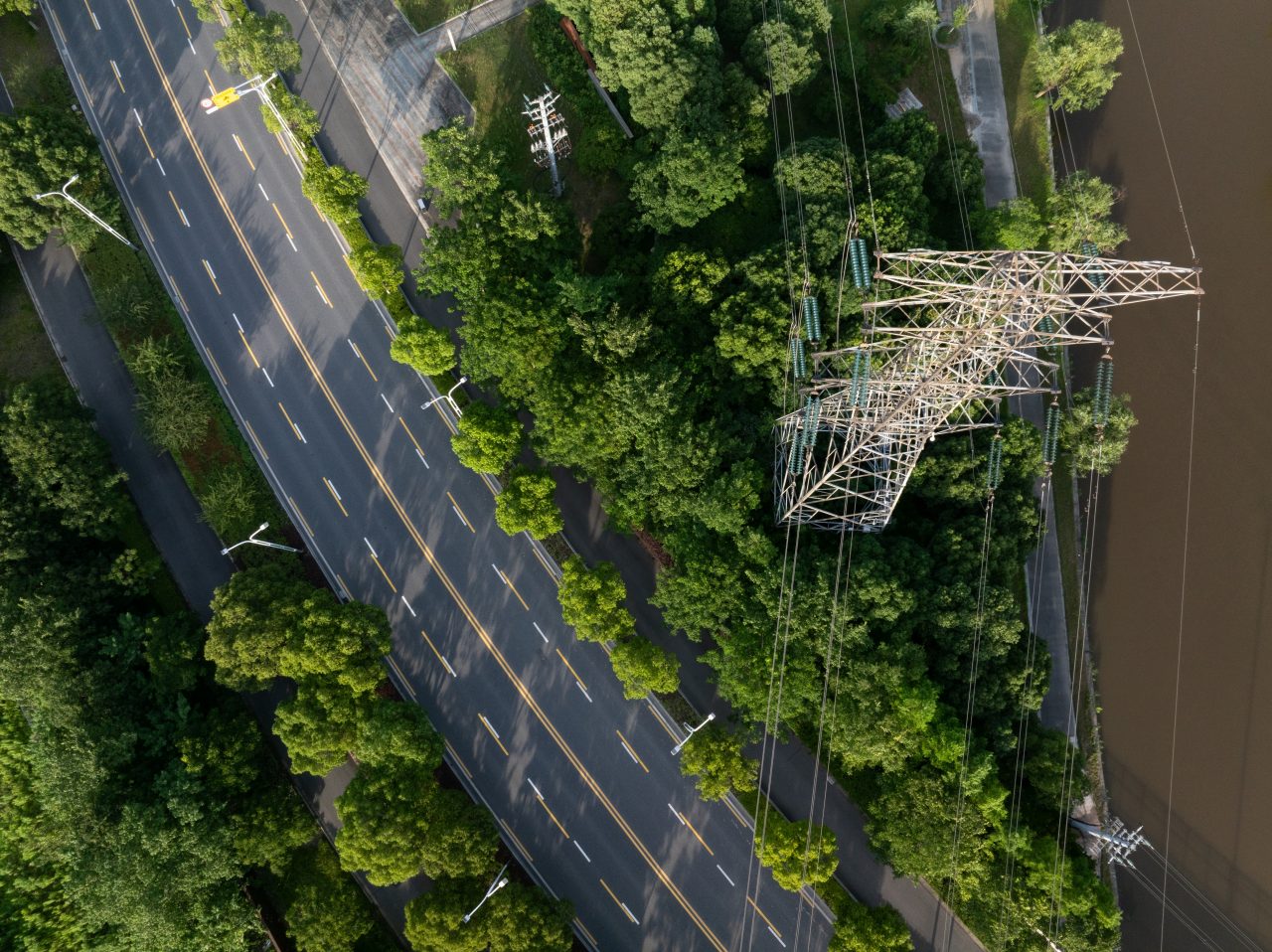 Aerial view of a power transmission tower surrounded by dense roadside vegetation near a highway, illustrating the importance of vegetation management for maintaining safe utility line operations.